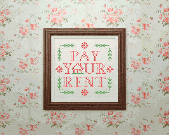 An embroidered picture of the words 'Pay your rent' in a frame, falling off a wall.